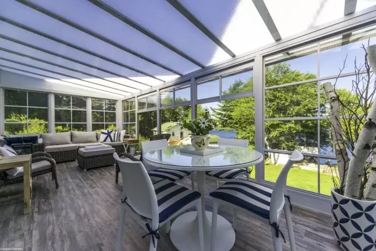 Sunroom interior featuring a round dining table with striped chairs, comfortable seating area, large windows, and a view of greenery, showcasing Sunspace By Sunrise Sunrooms' outdoor living solutions.