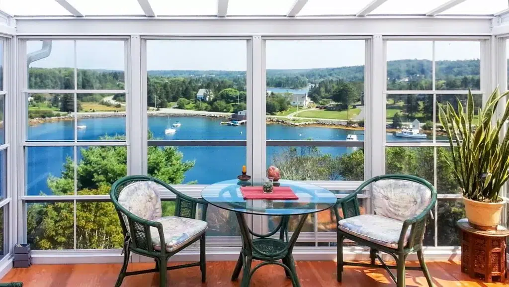 Sunroom with a glass table and two chairs overlooking a scenic waterfront view, featuring lush greenery and boats, ideal for relaxation and dining.