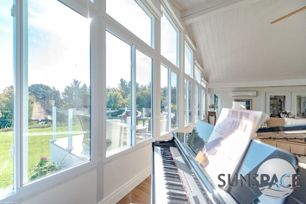 Sunroom interior with large windows showcasing natural light, featuring a piano and a view of the outdoor landscape, emphasizing a serene workspace for remote work and hobbies.