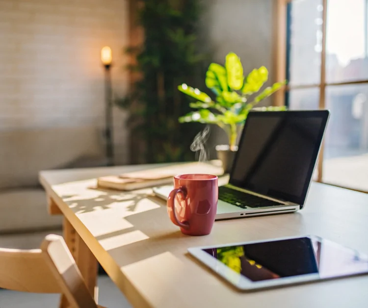 Cozy home office setup in a sunroom with a steaming mug, laptop, and greenery, emphasizing natural light and productivity.