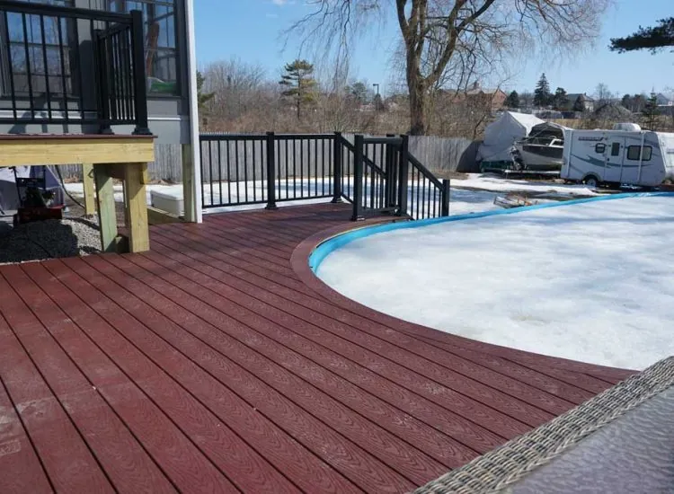 Red composite decking surrounding a pool area, featuring black railings and a view of a residential backyard, showcasing outdoor living space ideal for sunrooms and patio covers.